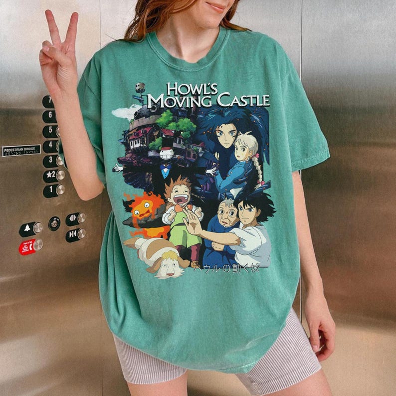 May include: A teal-colored t-shirt featuring a graphic from the animated film "Howl's Moving Castle." The design includes characters and the film's title. The shirt is worn by a person.