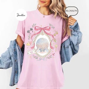 May include: A light pink t-shirt featuring a whimsical illustration of a large sphere within an oval frame, adorned with a pink bow, pearls, and floral accents. The design includes Mickey Mouse ears, a bunny, and a magic wand. The word "BLOSSOM" is in a speech bubble.