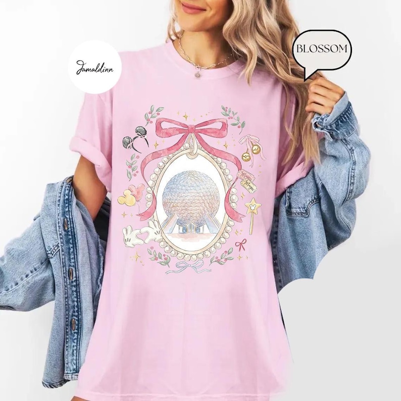 May include: A light pink t-shirt featuring a whimsical illustration of a large sphere within an oval frame, adorned with a pink bow, pearls, and floral accents. The design includes Mickey Mouse ears, a bunny, and a magic wand. The word "BLOSSOM" is in a speech bubble.