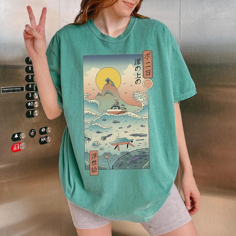 May include: Teal-colored t-shirt featuring a Japanese-inspired graphic. The graphic depicts a boat on a wave, a yellow sun, and Japanese text. The shirt is a relaxed fit.