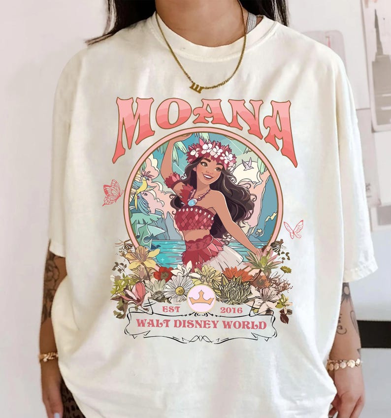 May include: Cream-colored t-shirt featuring a graphic of a woman in a floral crown, with the word "MOANA" in pink above her. The design includes flowers, butterflies, and the text "WALT DISNEY WORLD EST 2016".