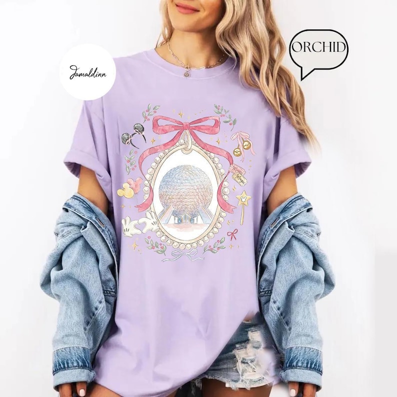 May include: A light purple t-shirt featuring a detailed illustration of a globe within an oval frame, adorned with a pink bow and floral accents. The shirt also includes Mickey Mouse ears, a magic wand, and the text "Jamaldinn" and "ORCHID".