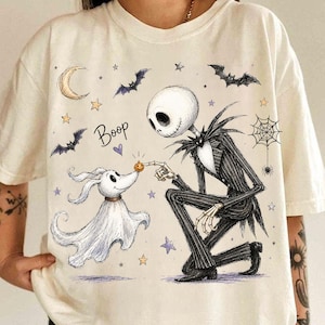 Könnte beinhalten: Ein cremefarbenes T-Shirt mit einer grafischen Illustration einer skelettartigen Figur im Nadelstreifenanzug, die vor einem Geisterhund kniet. Das Design zeigt einen Halbmond, Fledermäuse, Sterne, ein Spinnennetz, einen kleinen Kürbis und das Wort Boop.