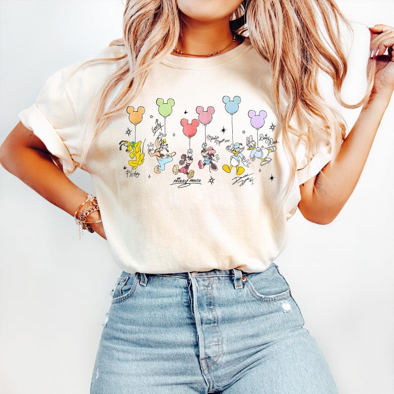 May include: Cream-colored t-shirt featuring a colorful graphic of Disney characters holding Mickey Mouse-shaped balloons. The characters include Pluto, Goofy, Mickey, Minnie, Donald, and Daisy Duck. The shirt has a relaxed fit.