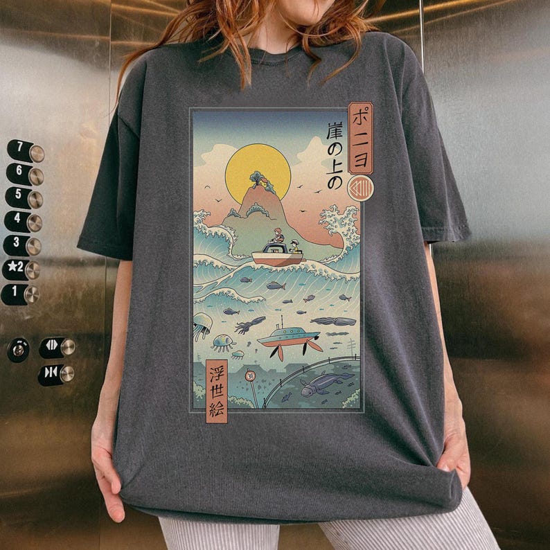 May include: Dark gray t-shirt featuring a Japanese-inspired illustration. The artwork depicts a boat on a wave, a submarine, and a rising sun. Japanese text is visible on the top right and bottom left of the graphic.