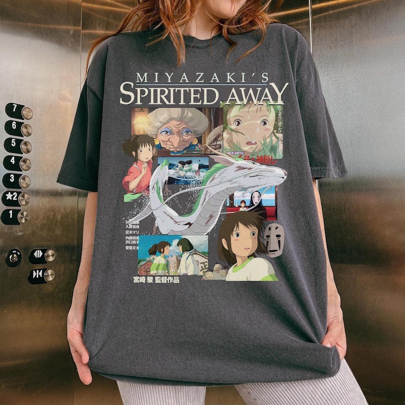 May include: Dark gray t-shirt featuring a colorful graphic from the animated film "Spirited Away." The design includes characters and the film's title. The shirt has short sleeves and a crew neck.