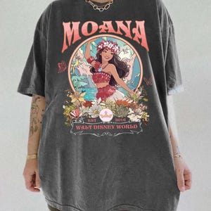 May include: Dark gray t-shirt featuring a graphic of Moana from Walt Disney World. The design includes the word "MOANA" in coral, a floral wreath, and the text "EST 2016 WALT DISNEY WORLD". The shirt has short sleeves and a relaxed fit.
