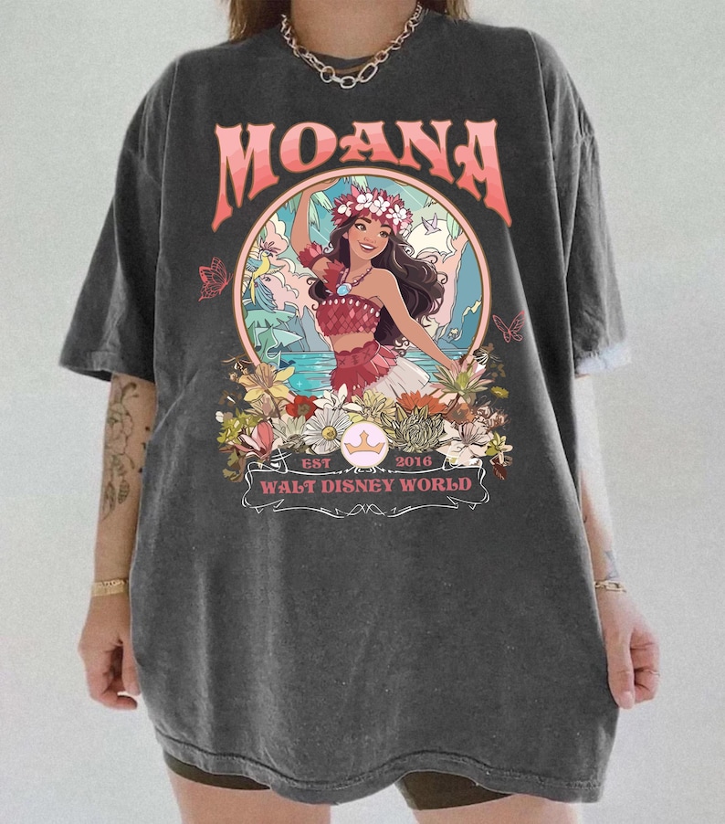 May include: Dark gray t-shirt featuring a graphic of Moana from Walt Disney World. The design includes the word "MOANA" in coral, a floral wreath, and the text "EST 2016 WALT DISNEY WORLD". The shirt has short sleeves and a relaxed fit.