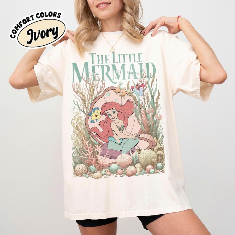 May include: Ivory-colored t-shirt featuring a graphic of Ariel from "The Little Mermaid" inside a seashell, surrounded by sea life. The text "The Little Mermaid" is above the graphic. The shirt has the words "Comfort Colors" and "Ivory" on the upper left.