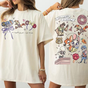 Puede incluir: Camiseta color crema con personajes de dibujos animados y texto. La parte delantera muestra personajes con diademas y la frase "You can be anything you wish to be." La parte trasera muestra m&aacute;s personajes y "The Big Donut."