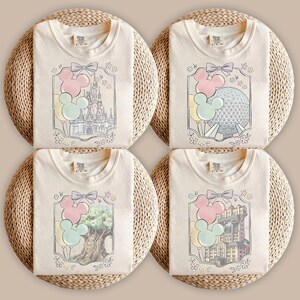Könnte beinhalten: Vier cremefarbene T-Shirts mit grafischen Drucken von Themenpark-Wahrzeichen, darunter ein Schloss, eine geodätische Kuppel, ein großer Baum und ein Hotel. Jedes Design enthält pastellfarbene Mickey-Ballons, eine Schleife und Blumen.