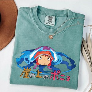May include: A seafoam green Comfort Colors t-shirt featuring a cartoon character with red hair and a blue and purple design. The Japanese text is printed below the character. A silver necklace with a pendant is draped across the shirt.