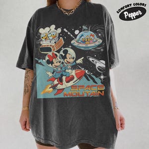 以下が含まれることがあります： 宇宙服を着た漫画のキャラクターがロケットや空飛ぶ円盤に乗っているグラフィックが描かれたチャコールグレーの半袖Tシャツ。SPACE MOUNTAINという文字が書かれています。デザインには星や惑星、COMFORT COLORS Pepperと書かれた小さな楕円形のラベルが含まれています。