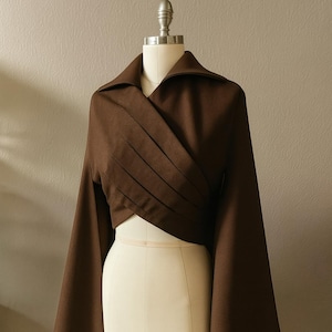 May include: A brown wrap top with long, flared sleeves. The top features a collared neckline and pleated detailing across the front. The garment is displayed on a mannequin, showcasing its design and fit. The fabric appears to be a medium-weight material.