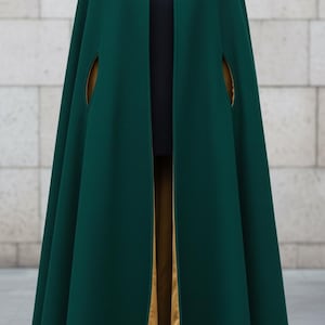 Può includere: Un mantello verde scuro con collo e chiusura decorativa. Il mantello presenta una fodera color oro visibile sull'apertura anteriore e negli spacchi delle braccia. L'indumento è esposto su un manichino su sfondo neutro.