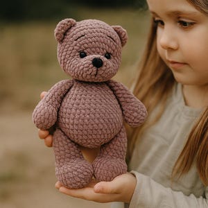 May include: A mauve-coloured, handmade teddy bear with a textured, crocheted appearance. The bear has a black nose and small, round eyes. The bear is being held by a person.