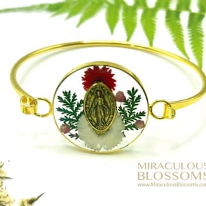 May include: Gold-tone bangle bracelet with a clear, round centerpiece. The centerpiece features a gold religious medallion, red and white flowers, and green foliage. The bracelet is adorned with the text "MIRACULOUS BLOSSOMS" and the website address.