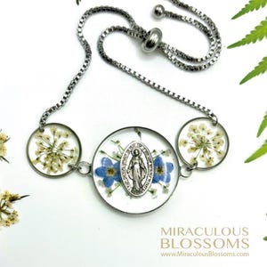 May include: A silver-toned adjustable bracelet featuring three clear, circular pendants. The pendants contain dried flowers and blue forget-me-nots. The center pendant holds a silver medallion of a religious figure. The text "MIRACULOUS BLOSSOMS" is visible.
