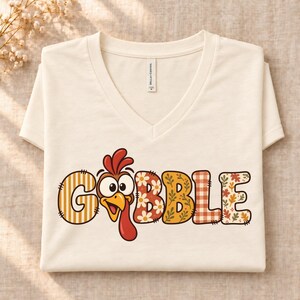 Può includere: Una t-shirt color crema con scollo a V che riporta la scritta GOBBLE, in cui la lettera O è sostituita da una testa di tacchino disegnata. Le lettere presentano vari motivi, tra cui righe, fiori bianchi, foglie autunnali e quadretti arancioni.