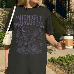 K&ouml;nnte beinhalten: Ein schwarzes Kurzarm-T-Shirt mit einem hellvioletten Grafikdruck, der den Text "MIDNIGHT MARGARITAS" &uuml;ber einer Illustration eines Margaritaglases, einer Mondsichel, Limettenscheiben, Agavenbl&auml;ttern und botanischen Elementen zeigt.