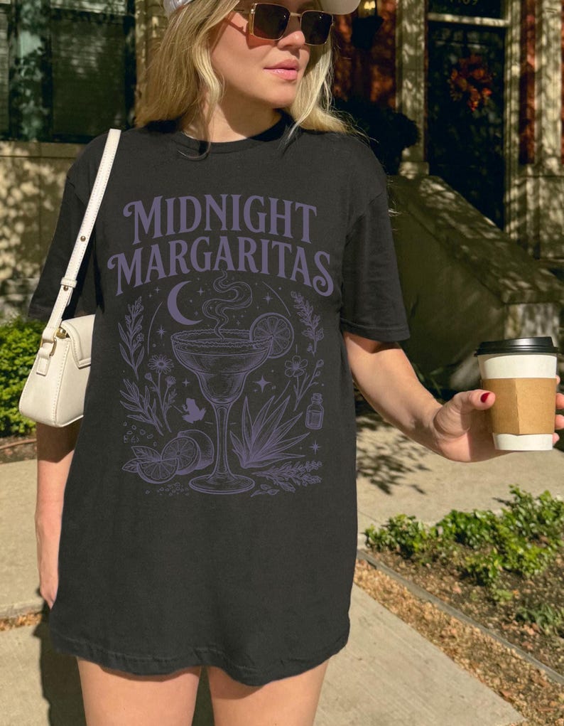 K&ouml;nnte beinhalten: Ein schwarzes Kurzarm-T-Shirt mit einem hellvioletten Grafikdruck, der den Text "MIDNIGHT MARGARITAS" &uuml;ber einer Illustration eines Margaritaglases, einer Mondsichel, Limettenscheiben, Agavenbl&auml;ttern und botanischen Elementen zeigt.