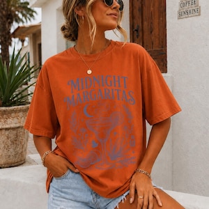 K&ouml;nnte beinhalten: Ein rostfarbenes Kurzarm-T-Shirt mit dem blauen Schriftzug "MIDNIGHT MARGARITAS" &uuml;ber einer grafischen Illustration eines Margaritaglases, einer Mondsichel und verschiedenen botanischen Elementen auf der Vorderseite des Kleidungsst&uuml;cks.