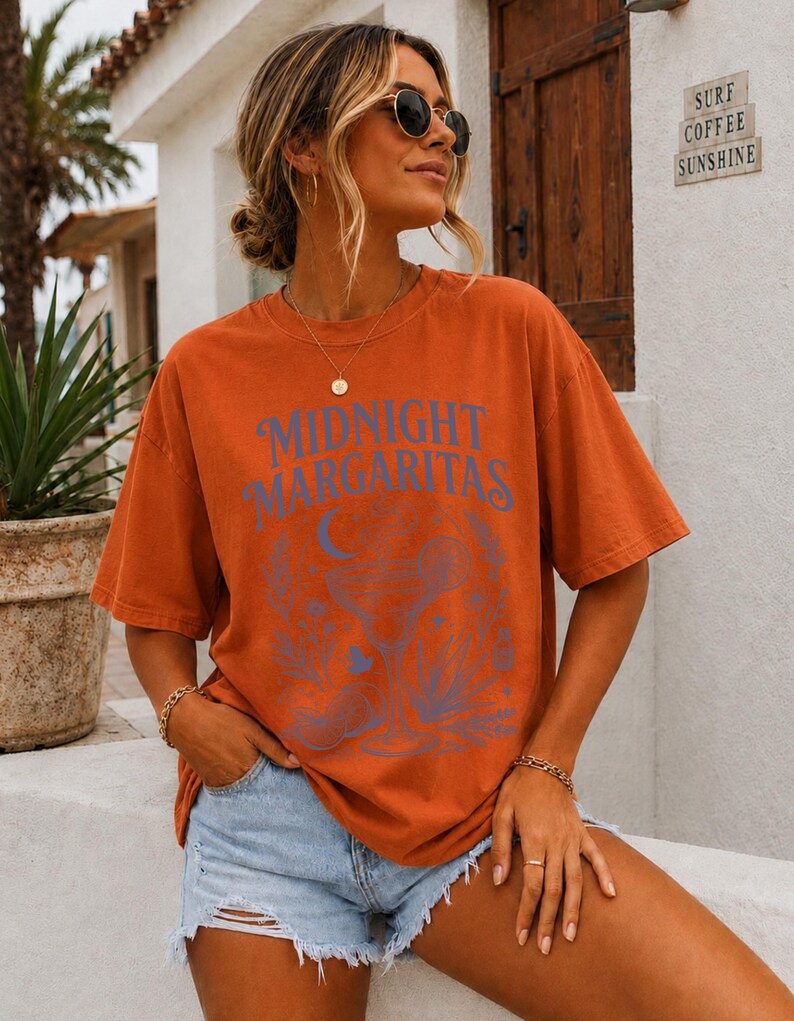 K&ouml;nnte beinhalten: Ein rostfarbenes Kurzarm-T-Shirt mit dem blauen Schriftzug "MIDNIGHT MARGARITAS" &uuml;ber einer grafischen Illustration eines Margaritaglases, einer Mondsichel und verschiedenen botanischen Elementen auf der Vorderseite des Kleidungsst&uuml;cks.