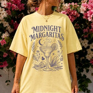 K&ouml;nnte beinhalten: Ein hellgelbes Kurzarm-T-Shirt mit einem dunkelblauen Grafikdruck. Das Design zeigt den Schriftzug "MIDNIGHT MARGARITAS" &uuml;ber einer Illustration eines Margaritaglases, einer Mondsichel, Sternen, Limettenscheiben und botanischen Elementen.