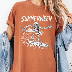 Puede incluir: Camiseta naranja quemado con la palabra "SUMMERWEEN" en blanco, sobre un esqueleto blanco con gafas de sol surfeando una ola. La camiseta tiene un corte holgado.