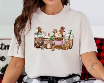 Puede incluir: Camiseta color crema con un gráfico festivo de cuatro bebidas temáticas navideñas. Las bebidas incluyen hombres de jengibre, casas de jengibre y otras decoraciones navideñas. La camiseta está hecha de un material suave.