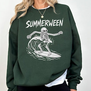 Könnte beinhalten: Dunkelgrünes Sweatshirt mit dem Wort "SUMMERWEEN" in weißer Schrift über einer weißen Grafik eines Skeletts, das eine Welle surft. Das Skelett trägt eine Sonnenbrille und reitet auf einem Surfbrett.