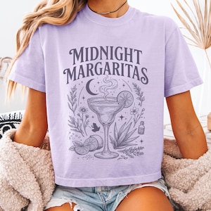 May include: A light purple short-sleeve t-shirt featuring a dark purple graphic print with the text "MIDNIGHT MARGARITAS" above an illustration of a margarita glass, a crescent moon, stars, lime slices, a small frog, and various botanical elements.