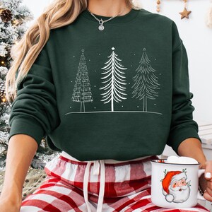 May include: A dark green sweatshirt featuring three white Christmas trees. The sweatshirt is paired with red and white plaid pajama pants. A white mug with a Santa Claus graphic is held in the hand.