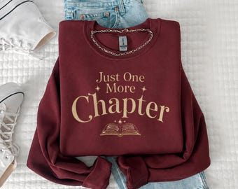 May include: A maroon long-sleeve crewneck sweatshirt featuring the gold text Just One More Chapter and a graphic of an open book with small stars. The sweatshirt is layered over light blue denim jeans and positioned near white canvas sneakers.