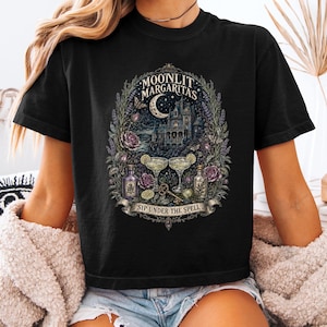May include: A black t-shirt featuring a detailed graphic print of a gothic castle under a crescent moon, two margarita glasses, bottles, flowers, and a key. The text reads "MOONLIT MARGARITAS" at the top and "SIP UNDER THE SPELL" on a banner at the bottom.