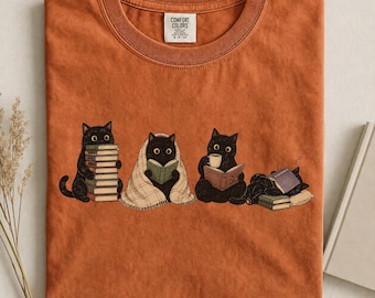 May include: A burnt orange crew neck t-shirt featuring a graphic of four black cats. One cat holds a stack of books, one is wrapped in a plaid blanket reading, one drinks from a mug while reading, and one sleeps on a book. The label reads Comfort Colors.