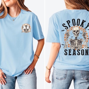 Puede incluir: Camiseta azul claro con un gr&aacute;fico de esqueleto. La parte delantera presenta un peque&ntilde;o dise&ntilde;o de esqueleto, mientras que la parte trasera muestra un esqueleto m&aacute;s grande con las palabras "Spooky Season" e ilustraciones de murci&eacute;lagos. Una prenda con tem&aacute;tica de Halloween.