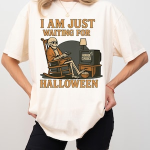 May include: Ivory t-shirt with a Halloween graphic. The design shows a skeleton in a rocking chair watching a vintage TV labelled "Horror Classics." Text reads "I AM JUST WAITING FOR HALLOWEEN."