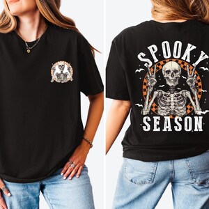 Puede incluir: Camiseta negra con un gr&aacute;fico de esqueleto. La parte delantera presenta un peque&ntilde;o dise&ntilde;o circular con dos esqueletos. La parte trasera muestra un dise&ntilde;o m&aacute;s grande con un esqueleto haciendo la se&ntilde;al de la paz, las palabras "Spooky Season" y murci&eacute;lagos.