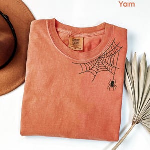 Pode incluir: Uma camiseta cor inhame com um desenho preto de teia de aranha e aranha. A camisa tem gola redonda e mangas curtas. A etiqueta diz "Comfort Colors". Um chapéu marrom e uma folha de palmeira seca estão no fundo.