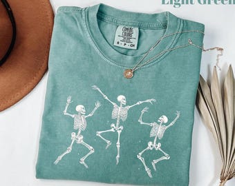 May include: Light green t-shirt with a crew neck and short sleeves. The shirt features a graphic of three dancing skeletons in white. A silver necklace with a pendant is draped across the shirt. The tag reads "Comfort Colors".