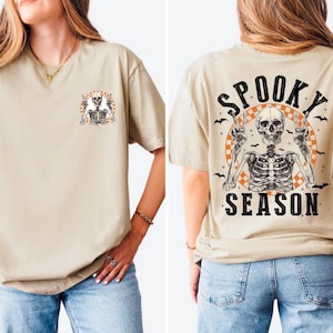 Puede incluir: Camiseta beige con un gr&aacute;fico de esqueleto en la parte delantera y trasera. La parte delantera presenta un peque&ntilde;o dise&ntilde;o de esqueleto, mientras que la parte trasera muestra un esqueleto m&aacute;s grande con las palabras "Spooky Season" e ilustraciones de murci&eacute;lagos. Prenda de vestir con tem&aacute;tica de Halloween.
