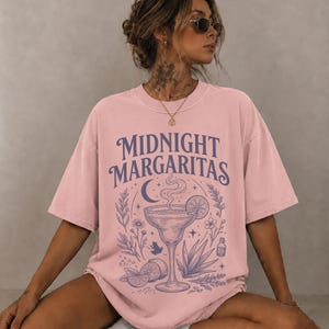 K&ouml;nnte beinhalten: Ein hellrosa T-Shirt mit kurzen &Auml;rmeln und einem dunkelvioletten Grafikdruck. Das Design zeigt den Text "MIDNIGHT MARGARITAS" &uuml;ber einer Illustration eines Margaritaglases, einer Mondsichel, Sternen, Blumen, einem kleinen Frosch und Limettenscheiben.