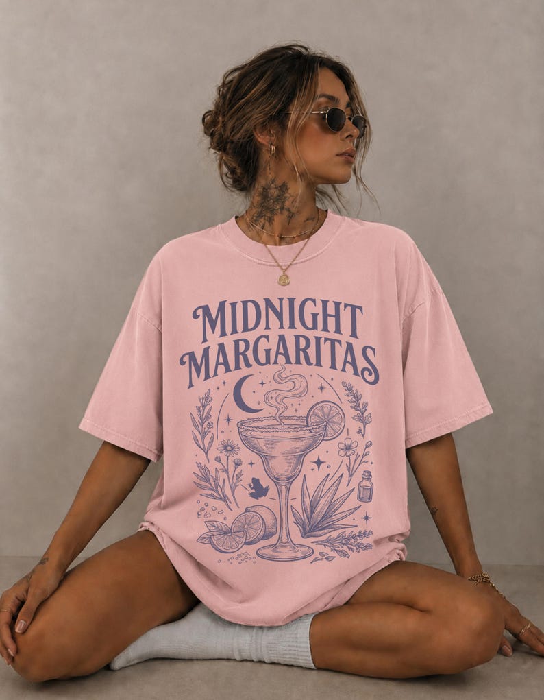 K&ouml;nnte beinhalten: Ein hellrosa T-Shirt mit kurzen &Auml;rmeln und einem dunkelvioletten Grafikdruck. Das Design zeigt den Text "MIDNIGHT MARGARITAS" &uuml;ber einer Illustration eines Margaritaglases, einer Mondsichel, Sternen, Blumen, einem kleinen Frosch und Limettenscheiben.