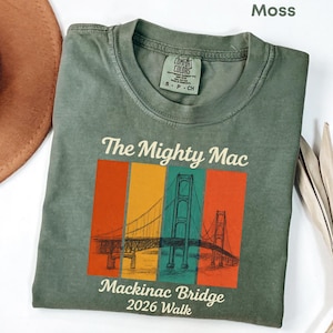 May include: A moss green t-shirt with a graphic of the Mackinac Bridge in a colorful rectangular design. The text "The Mighty Mac" and "Mackinac Bridge 2026 Walk" are printed on the shirt. The shirt is made of a soft material.