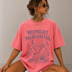 K&ouml;nnte beinhalten: Ein korallenfarbenes Kurzarm-T-Shirt mit einem dunkelblauen Grafikdruck. Das Design zeigt den Schriftzug "MIDNIGHT MARGARITAS" &uuml;ber einer Illustration eines Margaritaglases, einer Mondsichel, Sternen, Blumen und Agavenpflanzen.