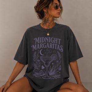 May include: A charcoal gray oversized t-shirt featuring the text "MIDNIGHT MARGARITAS" in a light purple font. The graphic includes a central margarita glass with steam, surrounded by illustrations of lime slices, agave plants, and stars.