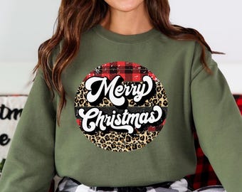 May include: Olive green sweatshirt with a circular graphic that says "Merry Christmas." The graphic has a red and black plaid pattern, a leopard print, and a black stripe. The text is in white script.