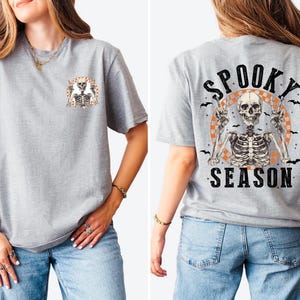 Puede incluir: Camiseta gris claro con un gr&aacute;fico de esqueleto. La parte delantera presenta un peque&ntilde;o dise&ntilde;o de esqueleto, mientras que la parte trasera muestra un esqueleto m&aacute;s grande con las palabras "Spooky Season" en negro. El dise&ntilde;o incluye acentos naranjas y negros.