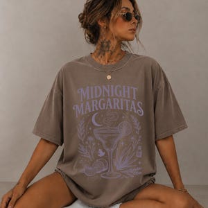 K&ouml;nnte beinhalten: Ein braunes Oversize-T-Shirt mit dem Schriftzug Midnight Margaritas in hellvioletter Schrift. Die Grafik zeigt ein zentrales Margaritaglas, eine Mondsichel, Zitrusfr&uuml;chte und botanische Elemente in einem passenden hellvioletten Farbton.